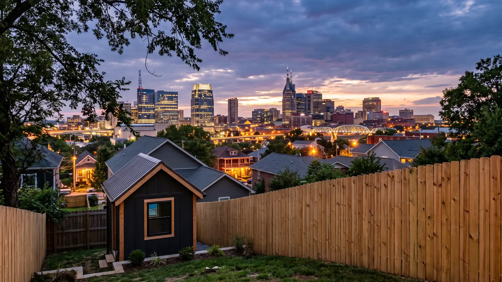 A dramatic dusk view of the Nashville skyline with warm lights over rooftops and a new shed and a wooden fence in the foreground