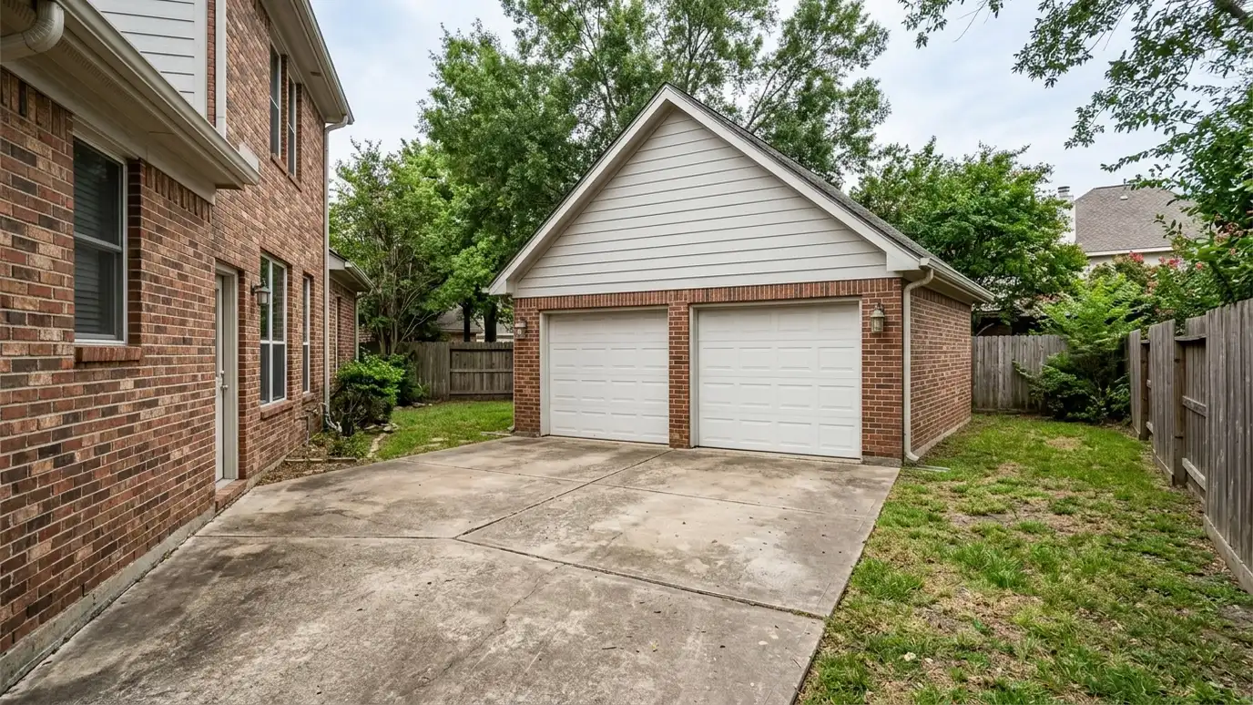 A detached two-car garage with brick and siding that match the house, white overhead doors and a gable roof at the end of a concrete driveway behind a brick home